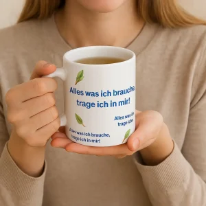 Eine Person hält eine weiße Tasse mit dem Text 'Alles was ich brauche, trage ich in mir!' und einem Blattmotiv. Die Person trägt einen beigen Pullover.